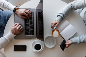 Overhead view of two people at a round white table, one typing on a laptop while the other holds a latte and phone with a notebook nearby.