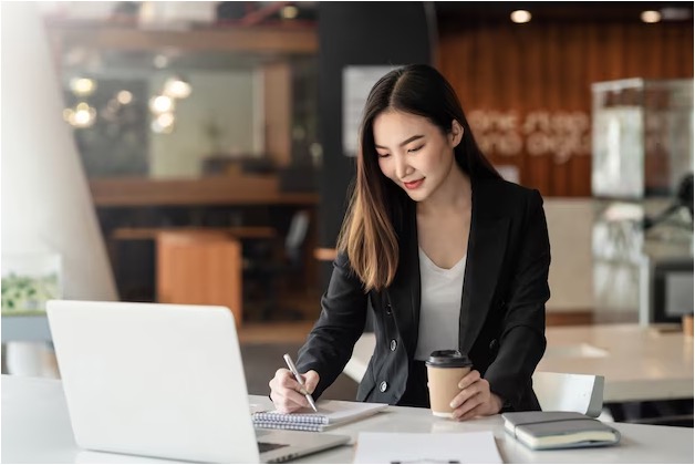 A businesswoman in a black blazer writing notes while holding a takeaway coffee cup at a desk with a laptop