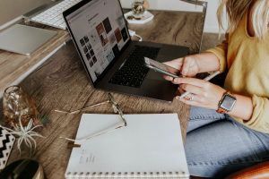 Writer drafting a journalistic-style article across a laptop, phone, and open notebook on a wooden desk