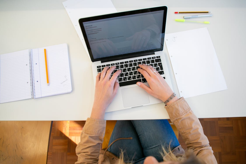 Person working on a laptop while drafting a press release, surrounded by a notepad, pencil, and printed pages.
