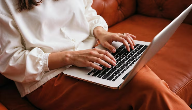 A person in a white shirt typing on a laptop while seated on an orange couch.