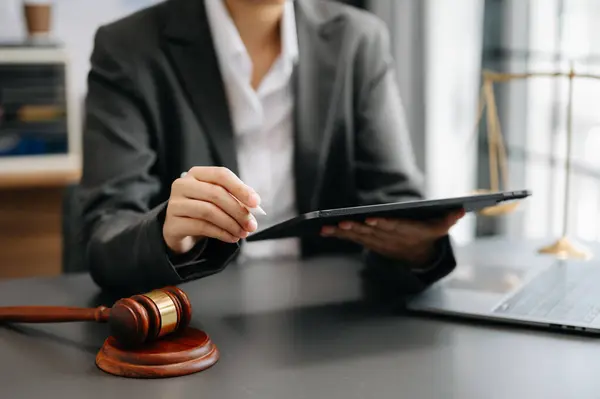A lawyer using a tablet with a stylus at a desk, with a gavel in the foreground and scales of justice in the background.