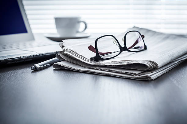 Reading glasses resting on a folded newspaper with a pen, laptop, and coffee cup on a desk.