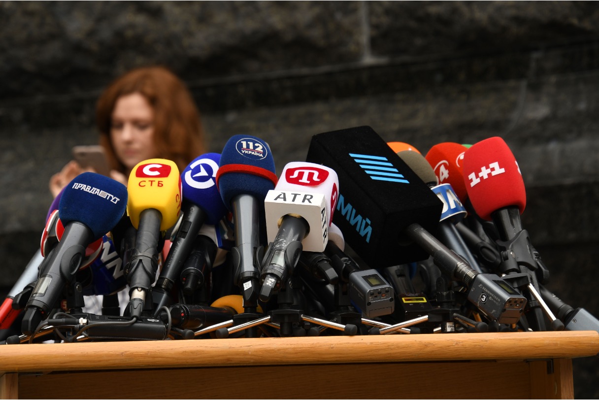 Multiple branded media microphones lined up on a press conference podium with a journalist in the background