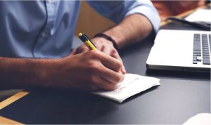 Person jotting handwritten notes on paper at a desk beside an open laptop