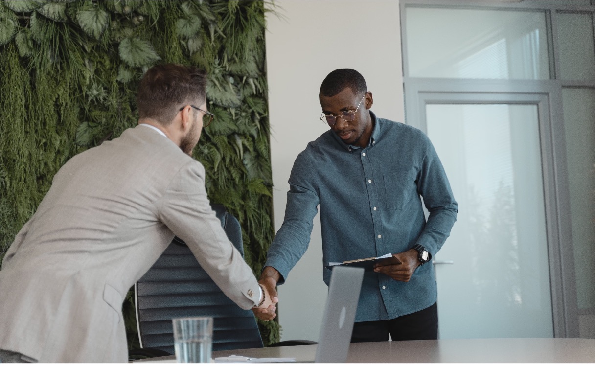 Two men shake hands across an office table with a laptop and a water glass