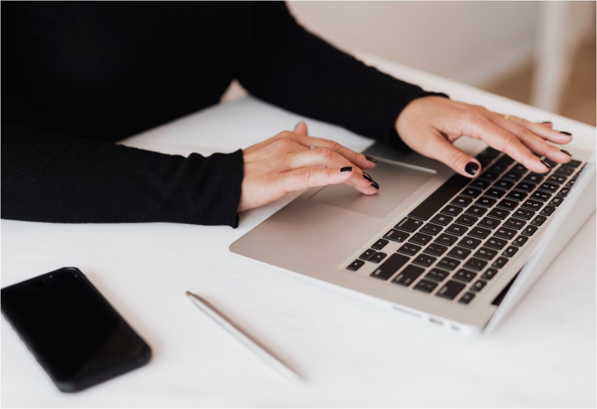 Woman in black typing on a silver laptop at a white desk with a phone and pen